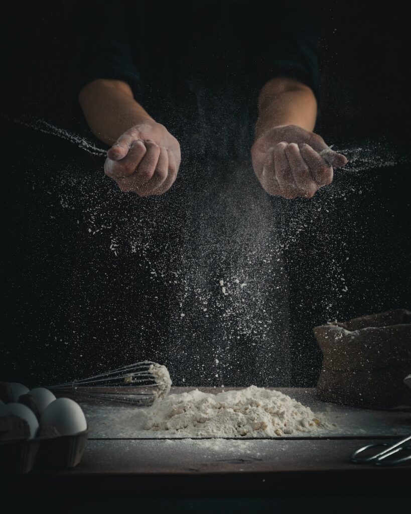 person pouring flour on table beside eggs and whisk